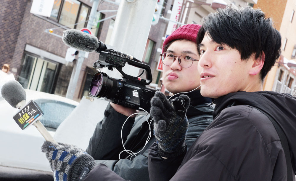 地下鉄の駅前で街の人に声をかける「ナイス街録ちゃん」のロケ現場。 カメラを構えるのは日向寺駿也さん、マイクを手にするのが太田夏主馬さん。